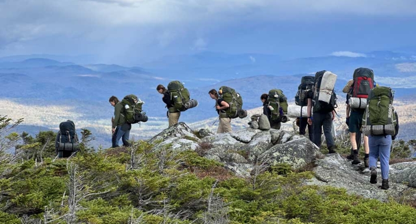 A group of students wearing backpacks hike along a rocky path at high elevation. Far below them lies more mountainous terrain.